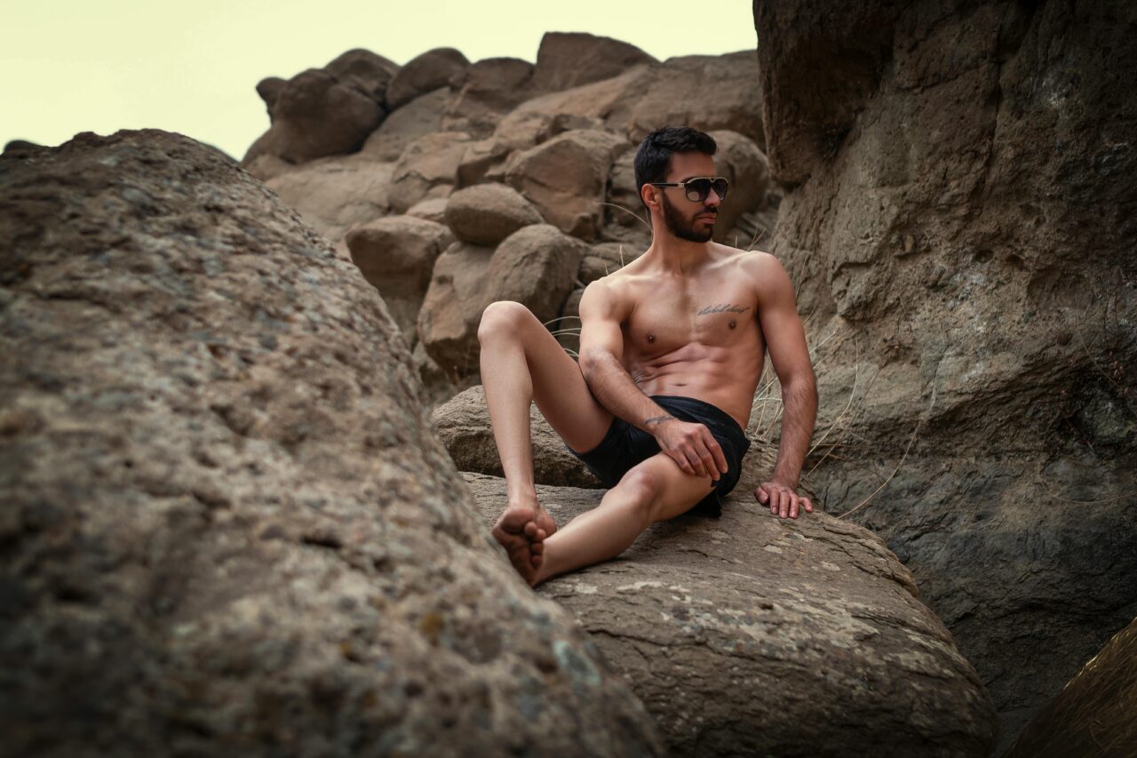 A shirtless man in shorts sits confidently on rocky terrain, embracing the wild outdoors.