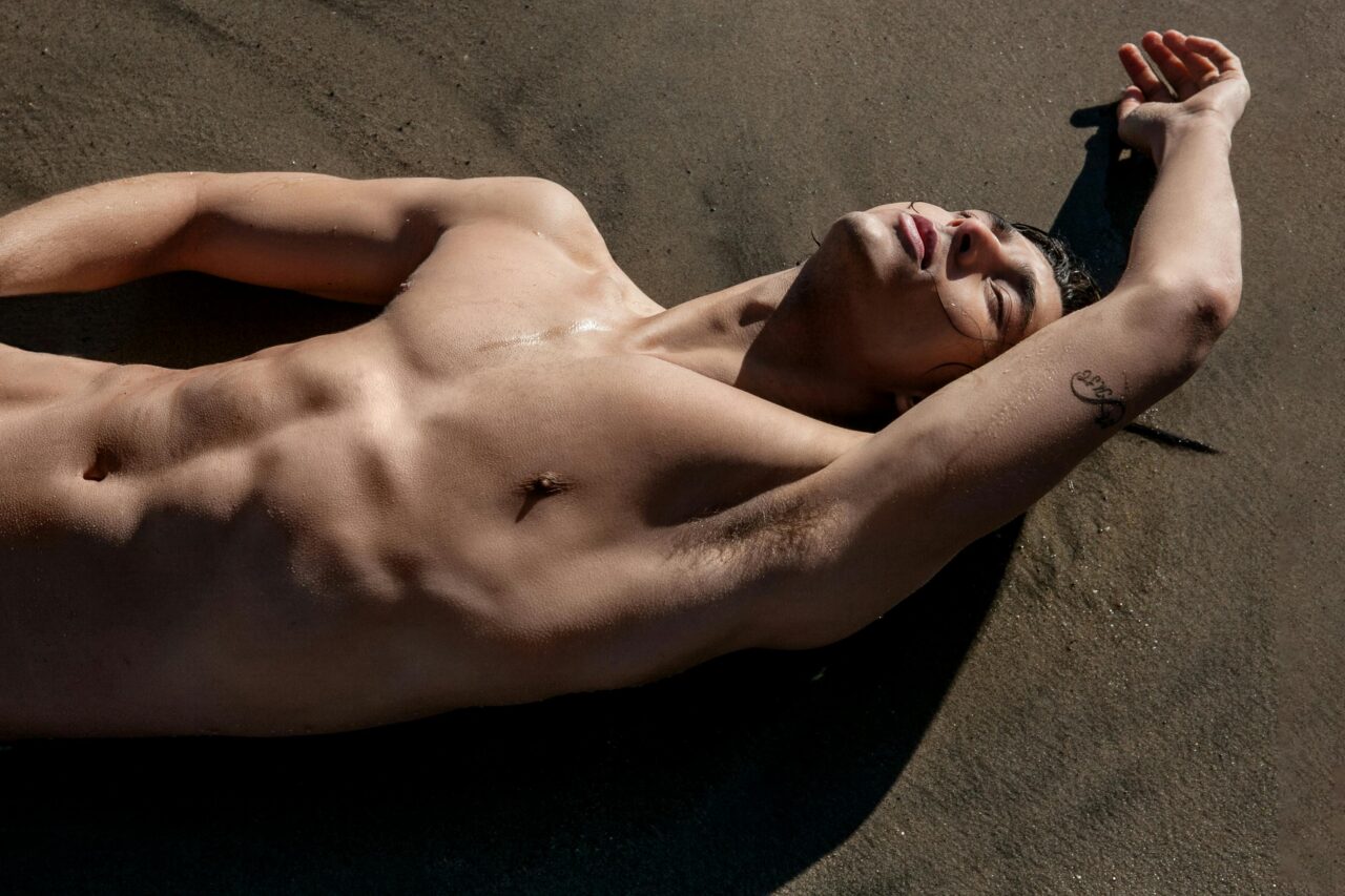 A muscular man sunbathing shirtless on the sandy beach with a tattoo visible.