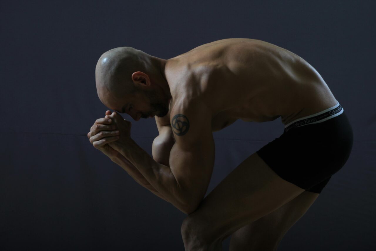 Bald, muscular man bending and posing shirtless against a gray background.