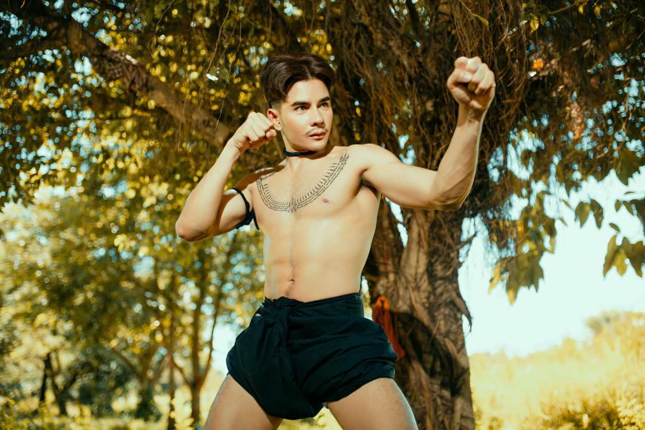 Topless man with tattoos posing in a boxing stance outdoors under a tree.