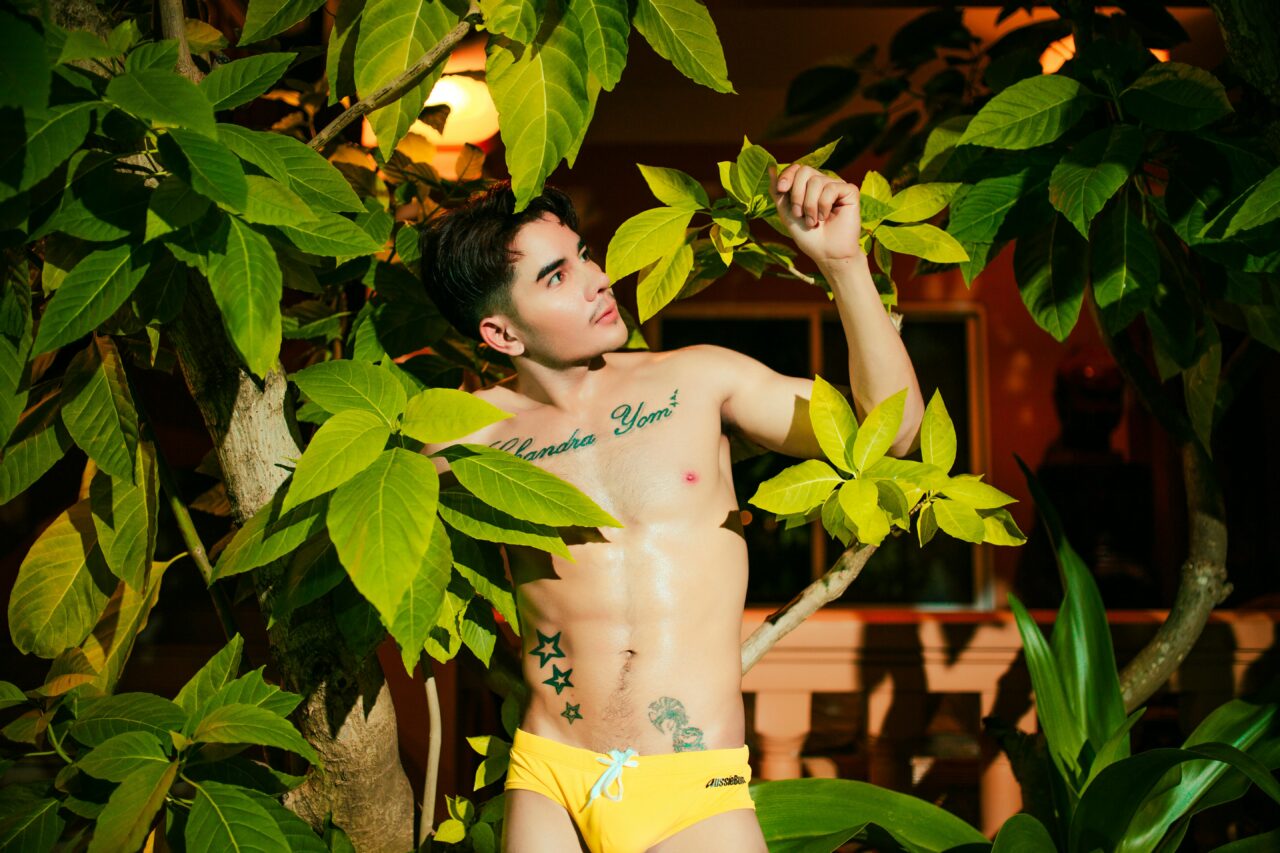 Portrait of a young man in yellow swimwear standing among tropical leaves at night.
