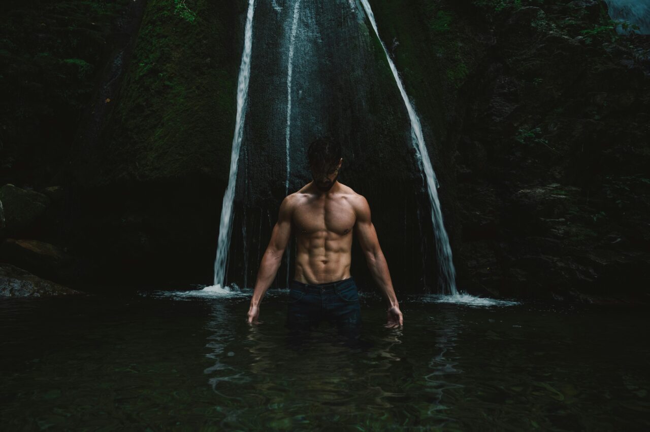 A shirtless man stands in water, showcasing muscles under a mystical waterfall.