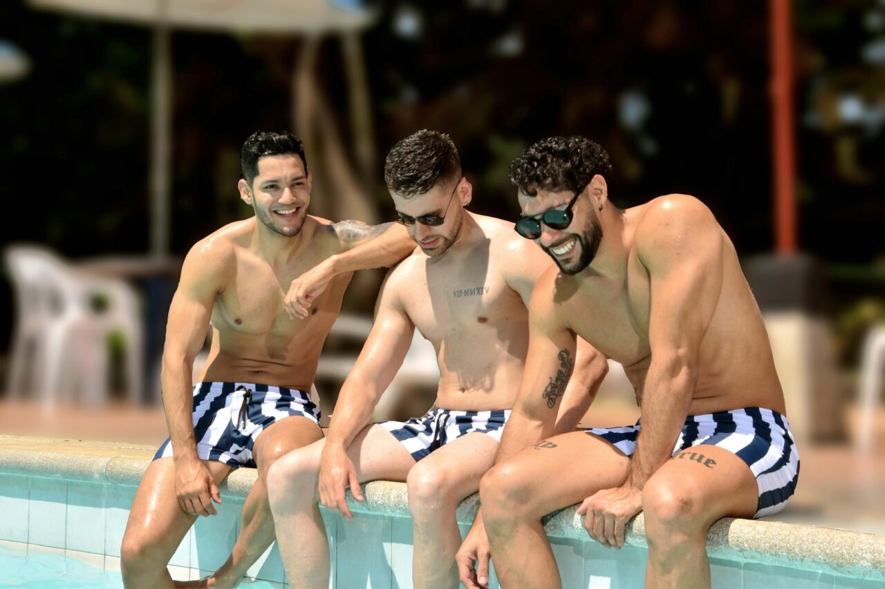 Three young men in striped swim trunks enjoying a sunny day by the pool, smiling and relaxed.