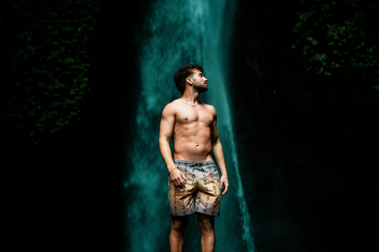 A young man stands confidently before a majestic waterfall in Bali's lush outdoors.