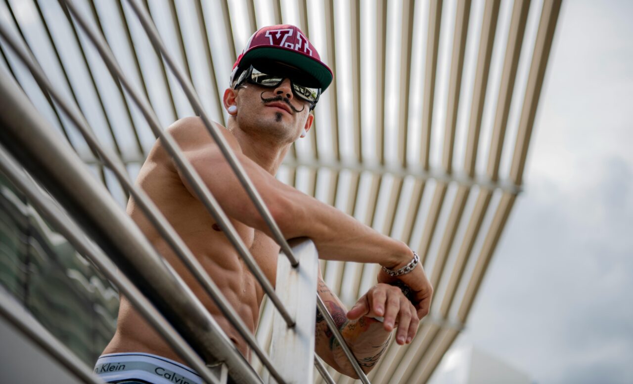 Shirtless man with tattoos and sunglasses casually leaning on a balcony with modern architecture.