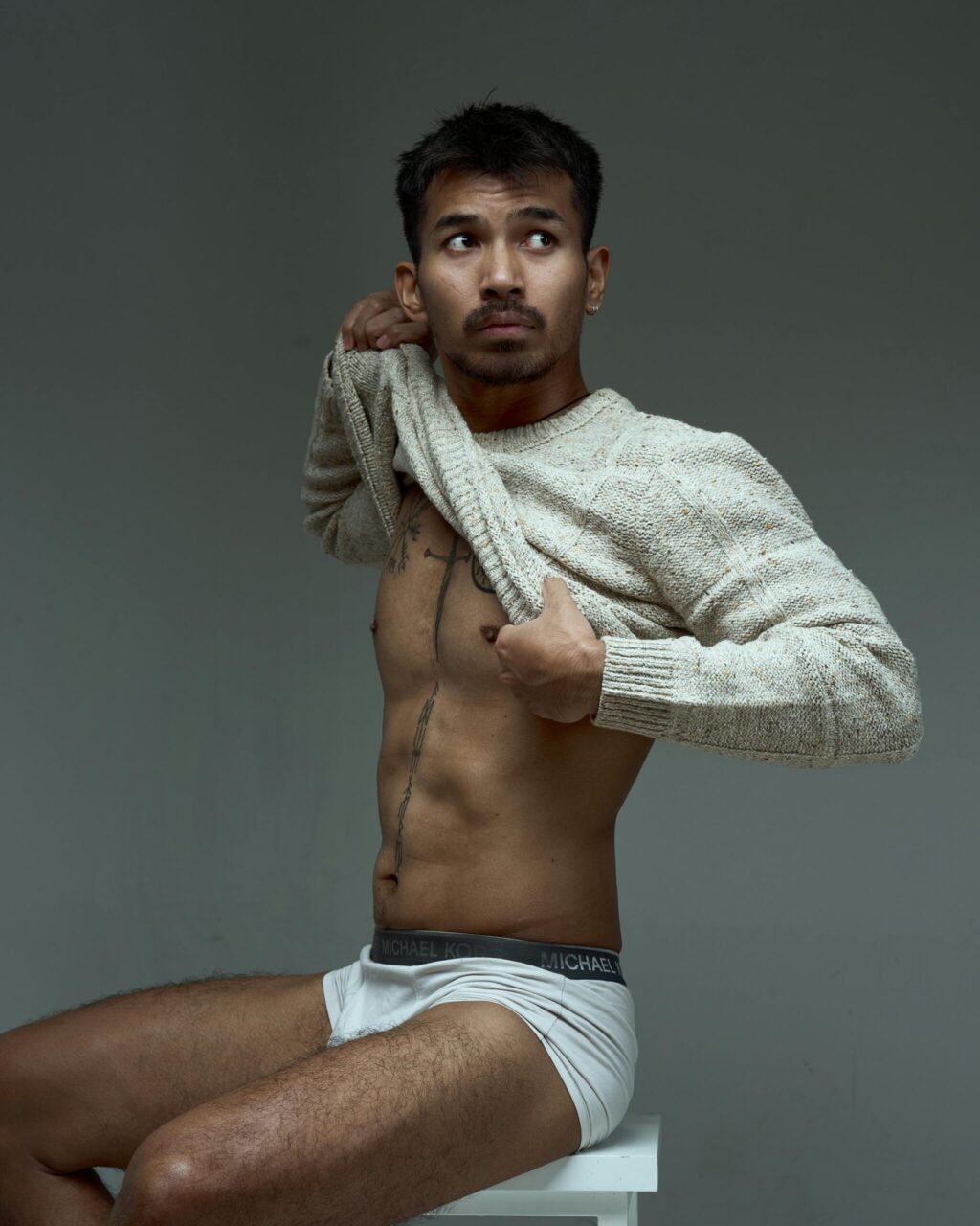 Stylish portrait of a shirtless man in a casual sweater against a gray background.