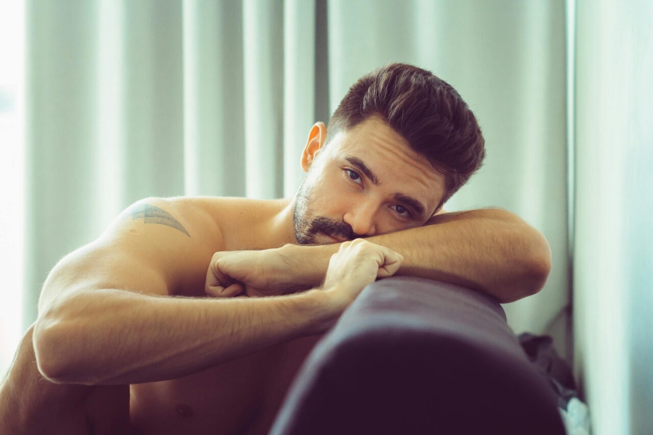 A thoughtful shirtless man with a beard lounging comfortably indoors, conveying a relaxed mood.