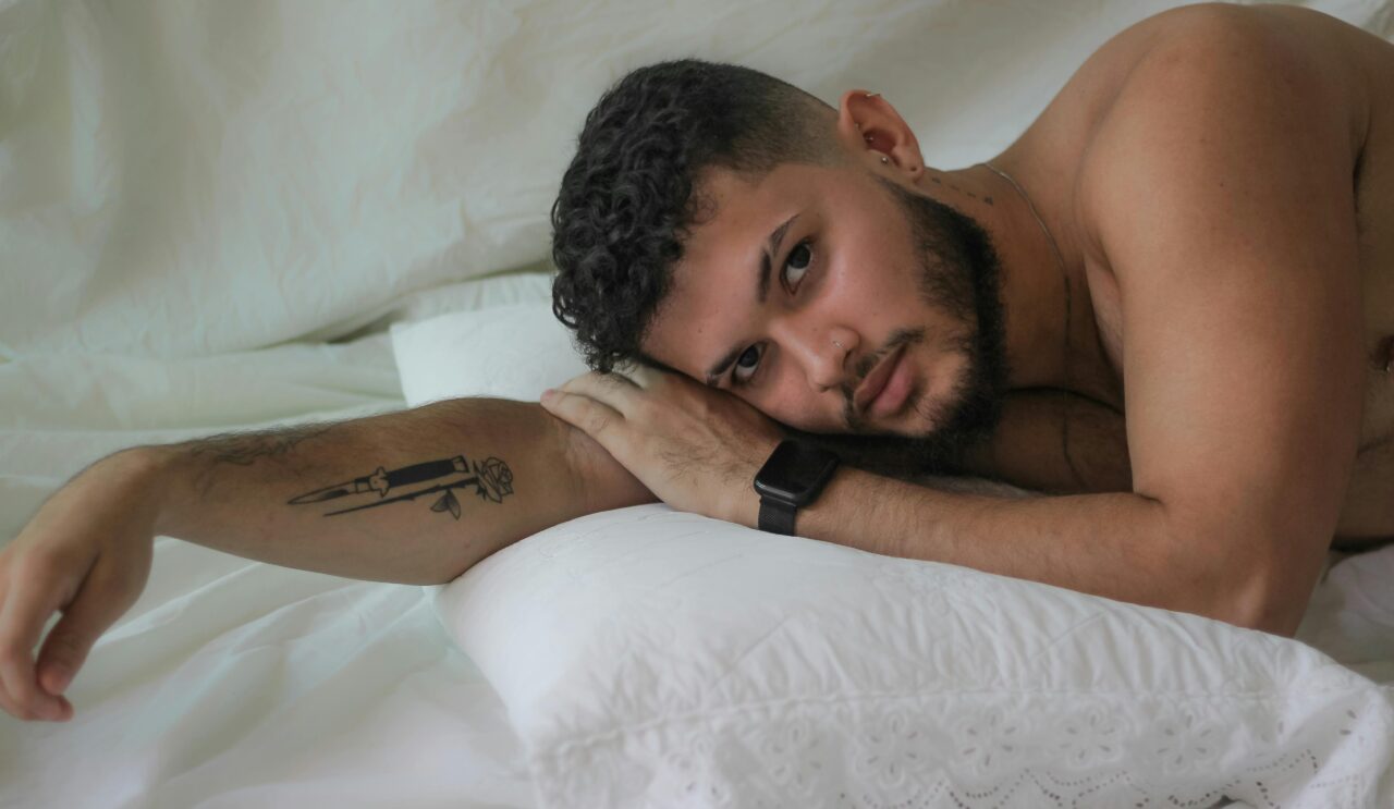 A bearded man with a tattoo reclines comfortably on white pillows, exuding serenity and style.