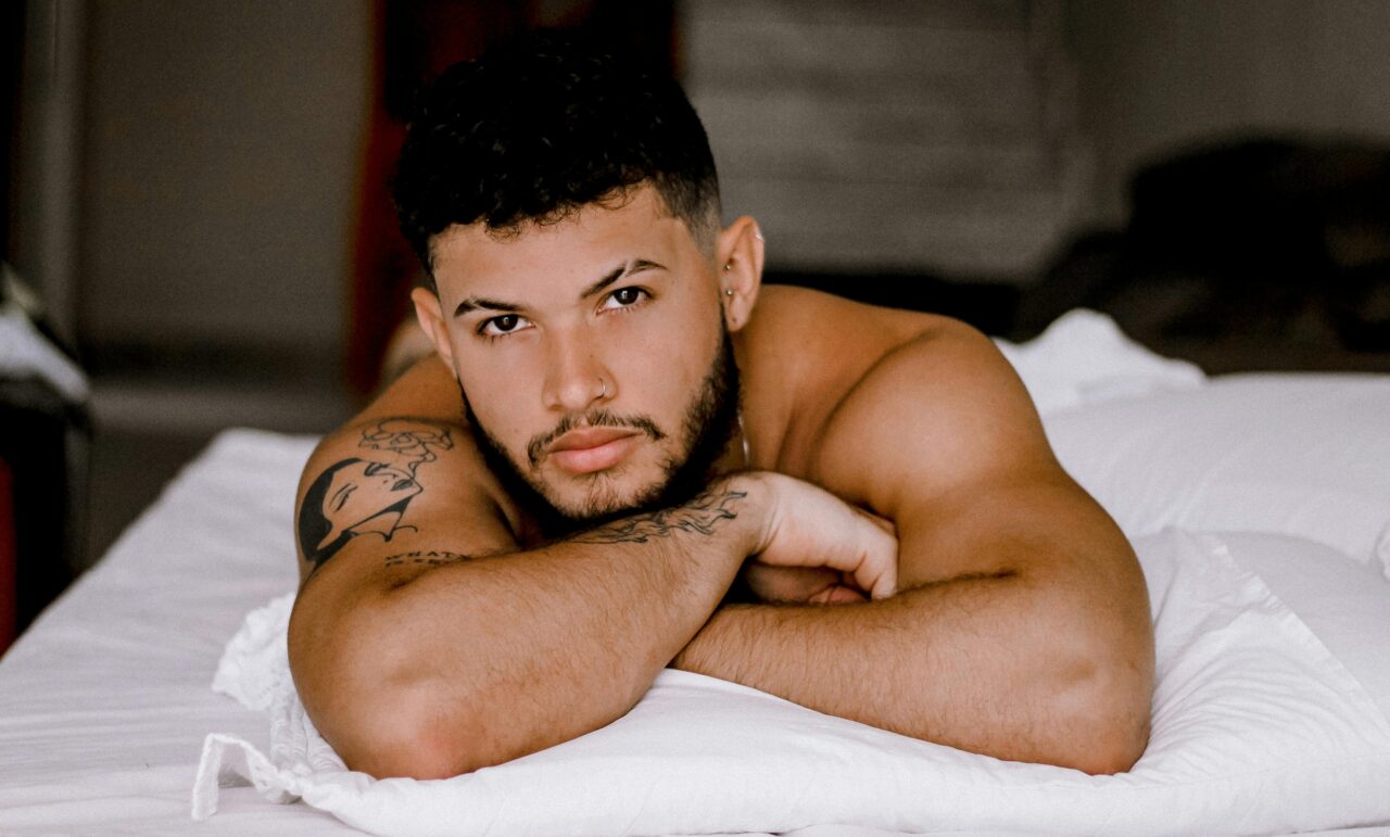 A bearded man with tattoos is resting on a white bed, exuding a relaxed and contemplative mood.