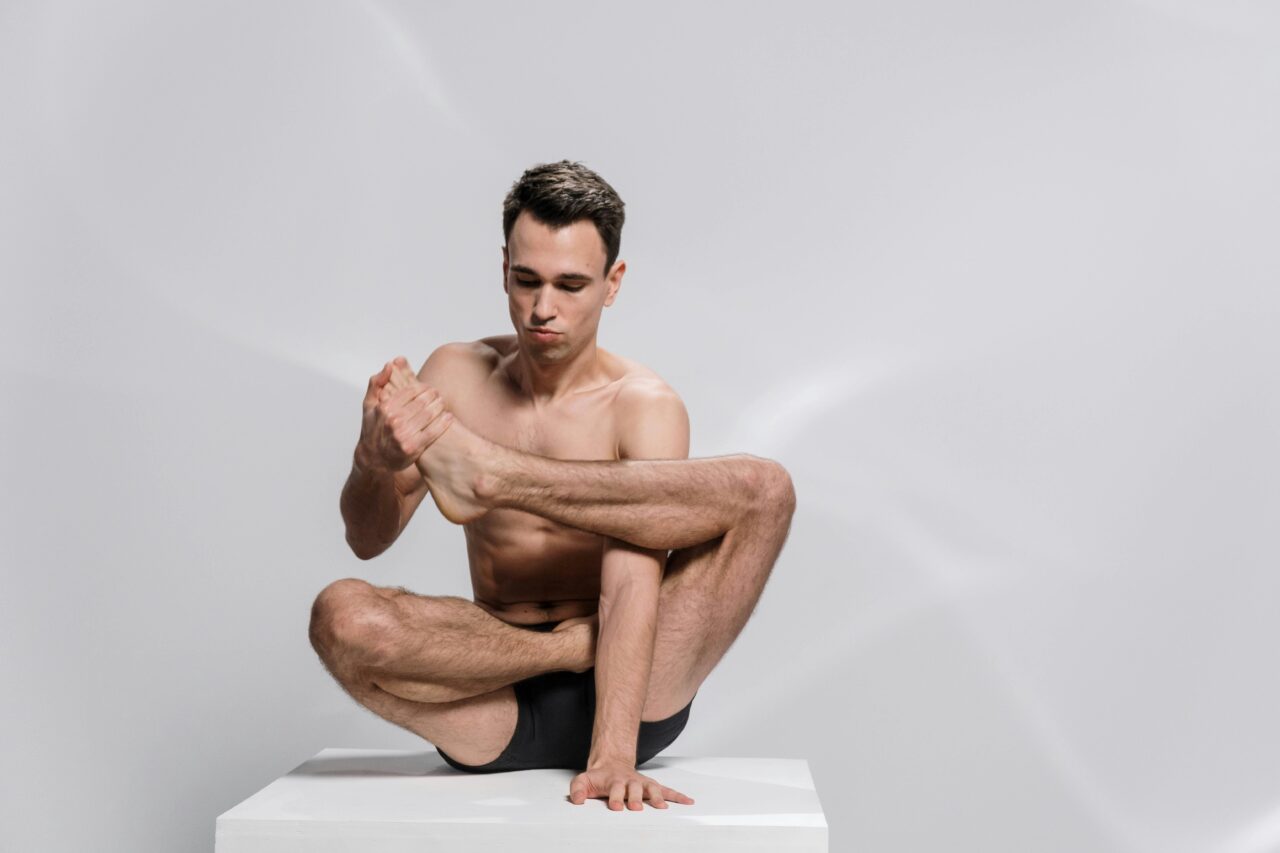Shirtless man demonstrating flexibility in a seated yoga pose on a white background.