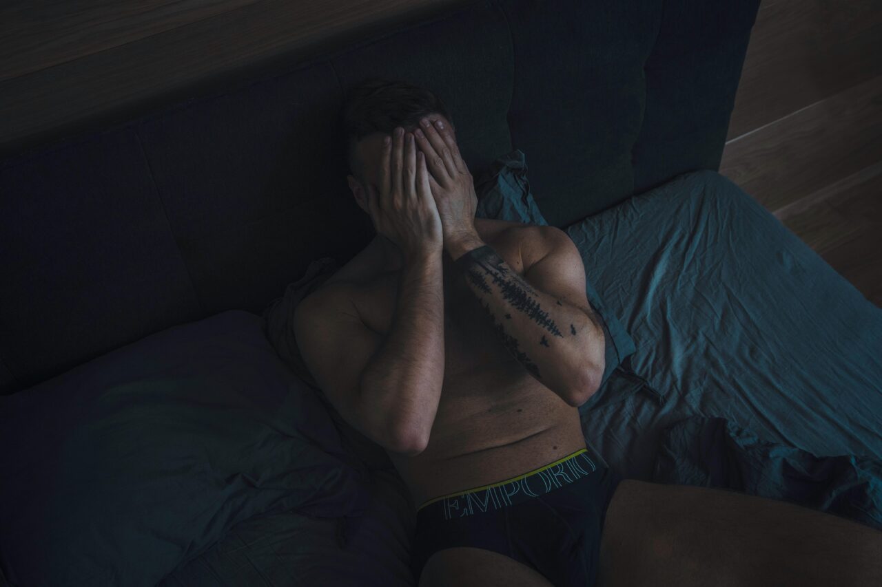 A shirtless man sits on a bed indoors, covering his face with his hands.
